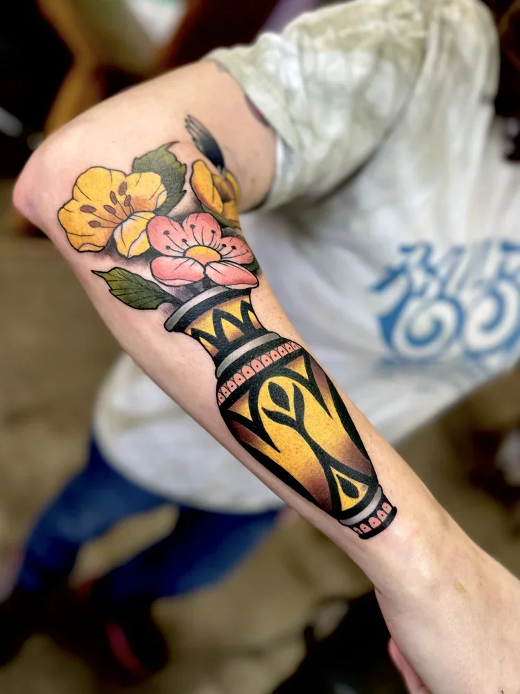 Close-up of a colorful tattoo on a person's forearm featuring a floral arrangement of yellow and pink flowers with green leaves, and an orange and black tribal-style design vase.