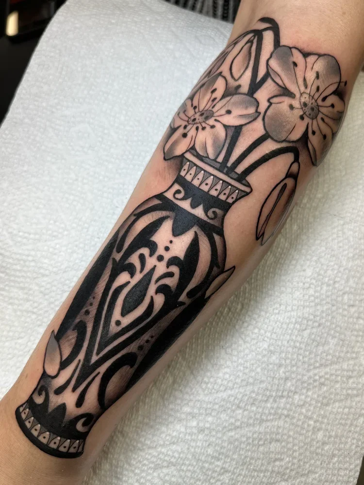 Tattoo of a decorative black vase with flowers on a person's forearm.
