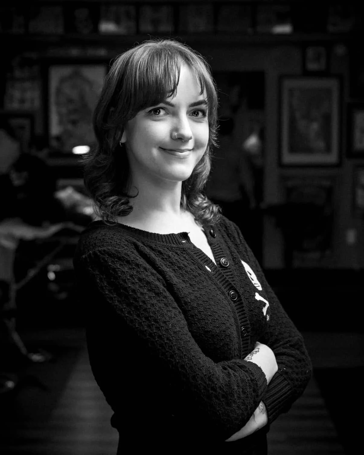 Black and white portrait of a young woman with shoulder-length wavy hair and a nose piercing, smiling with arms crossed, in a room with framed pictures on the wall - Hannah Turner, tattoo artist.