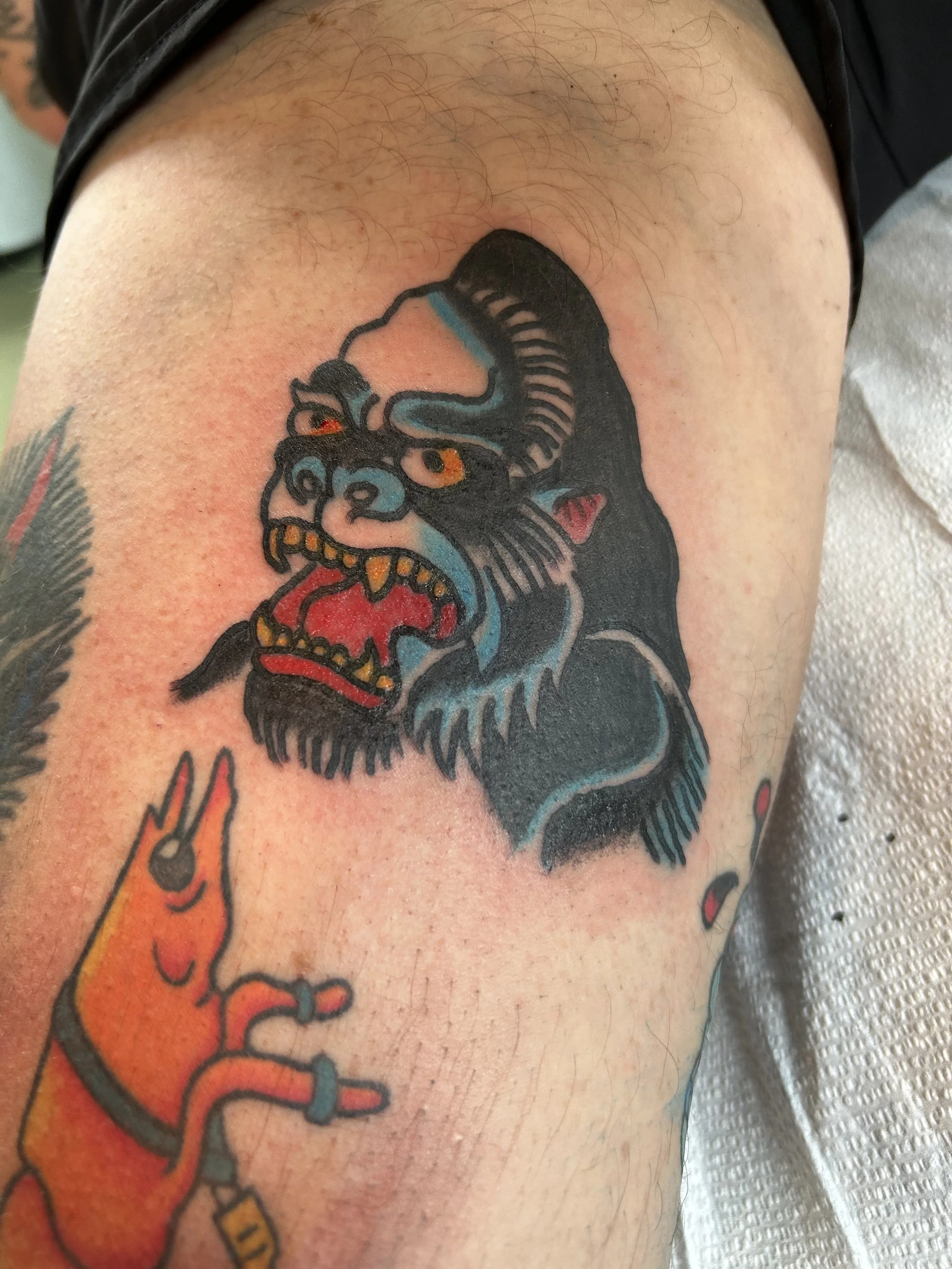 Tattoo of a fierce, traditional-style mythological creature with orange eyes, horns, sharp teeth, and fangs, depicted in black, blue, red, and yellow ink on the person's upper arm.