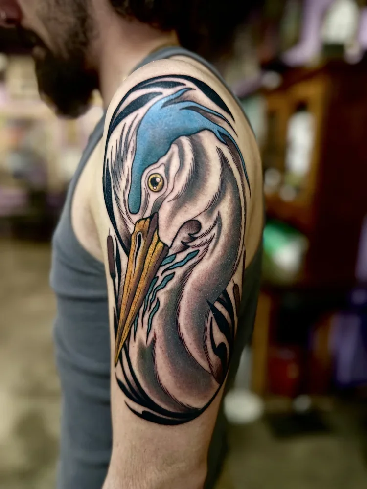 Colorful tattoo of a heron on a person's upper arm, featuring blue, white, black, and brown details.