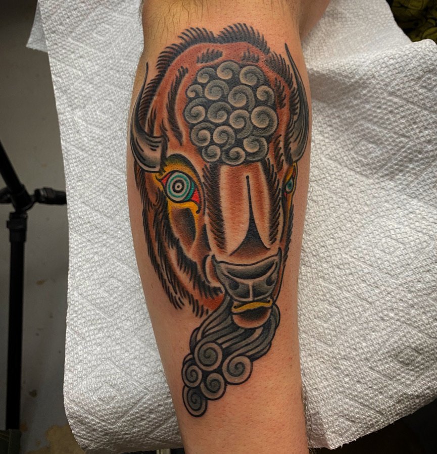 Tattoo of a bear with colorful eyes, a stylized nose, and swirling smoke or fur design on the arm.