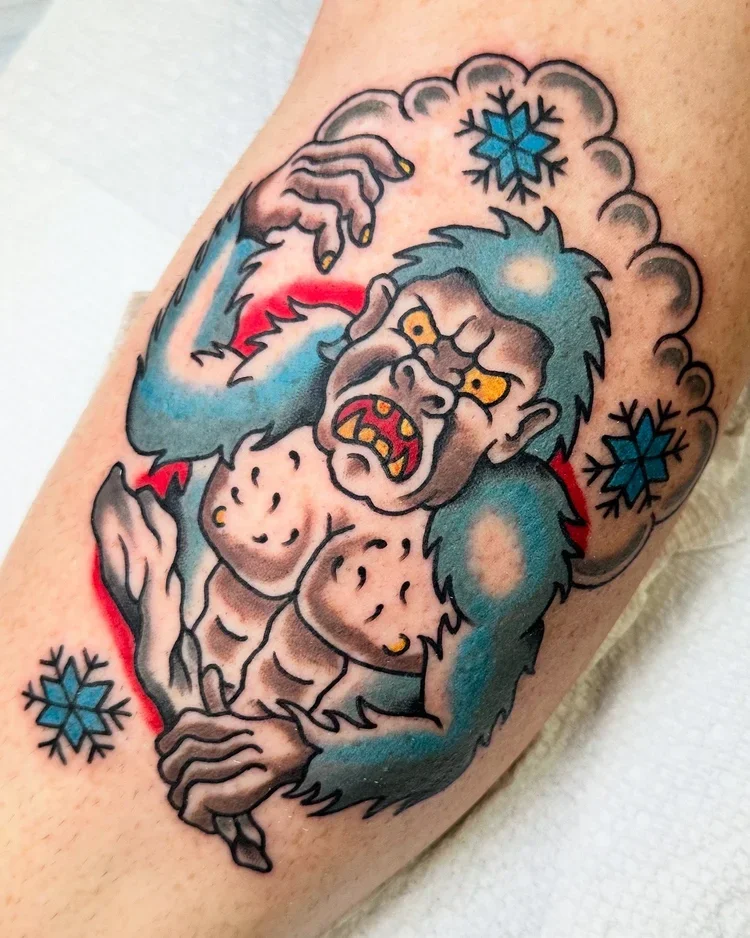 Tattoo of a comic-style Yetti with yellow eyes, long blue hair, large sharp teeth, and angry expression. It is holding a piece of ice surrounded by snowflakes.