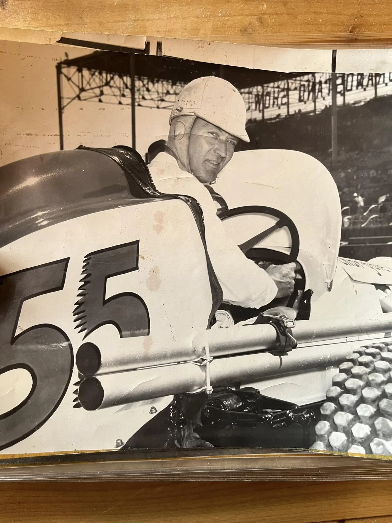 Black and white photograph of a race car driver wearing a helmet, sitting in his open-cockpit race car numbered 55, at a racetrack with grandstands in the background.
