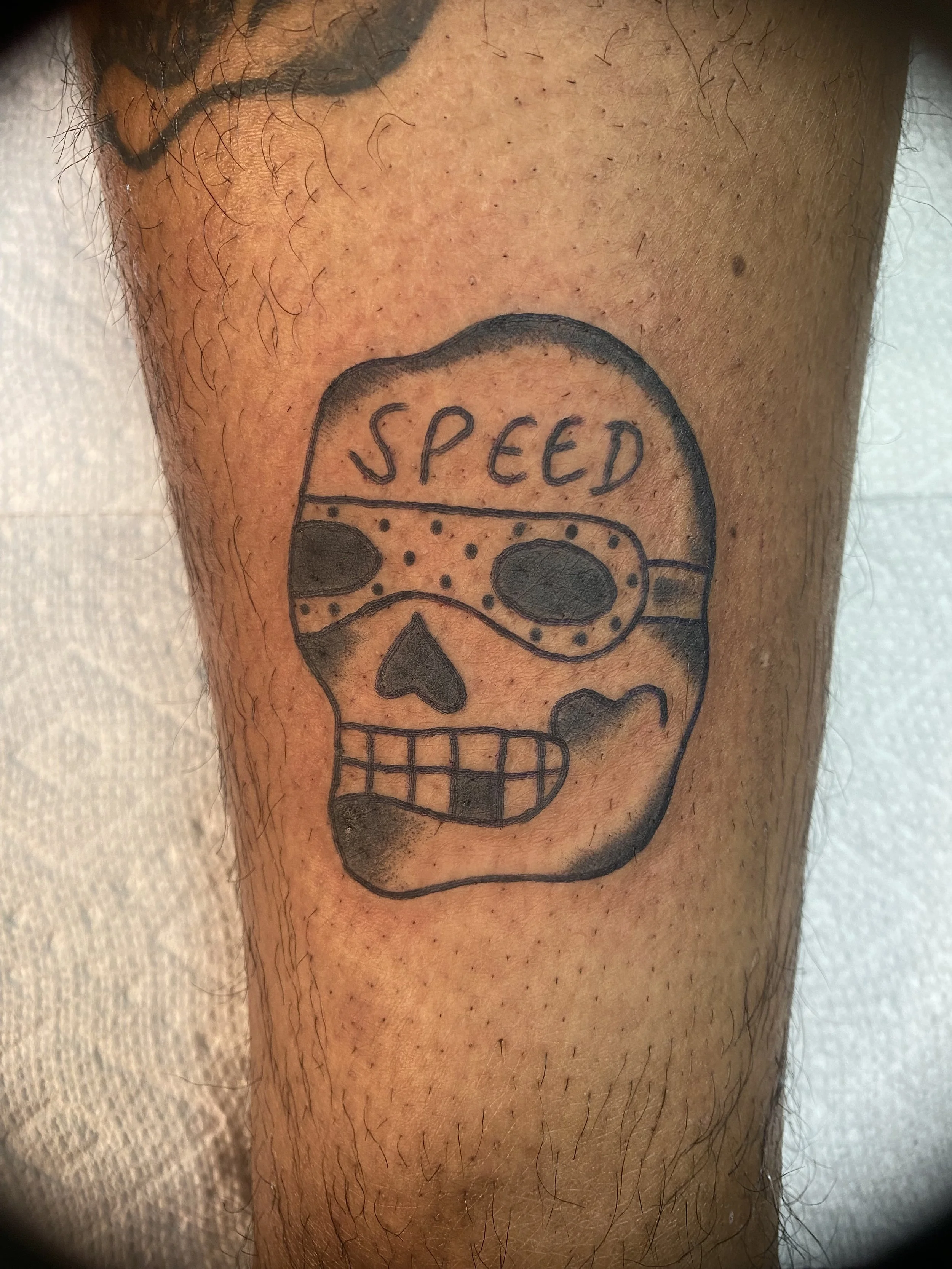 Tattoo of a skull with sunglasses, the word 'SPEED' on its forehead, and a missing eye, on a person’s leg.
