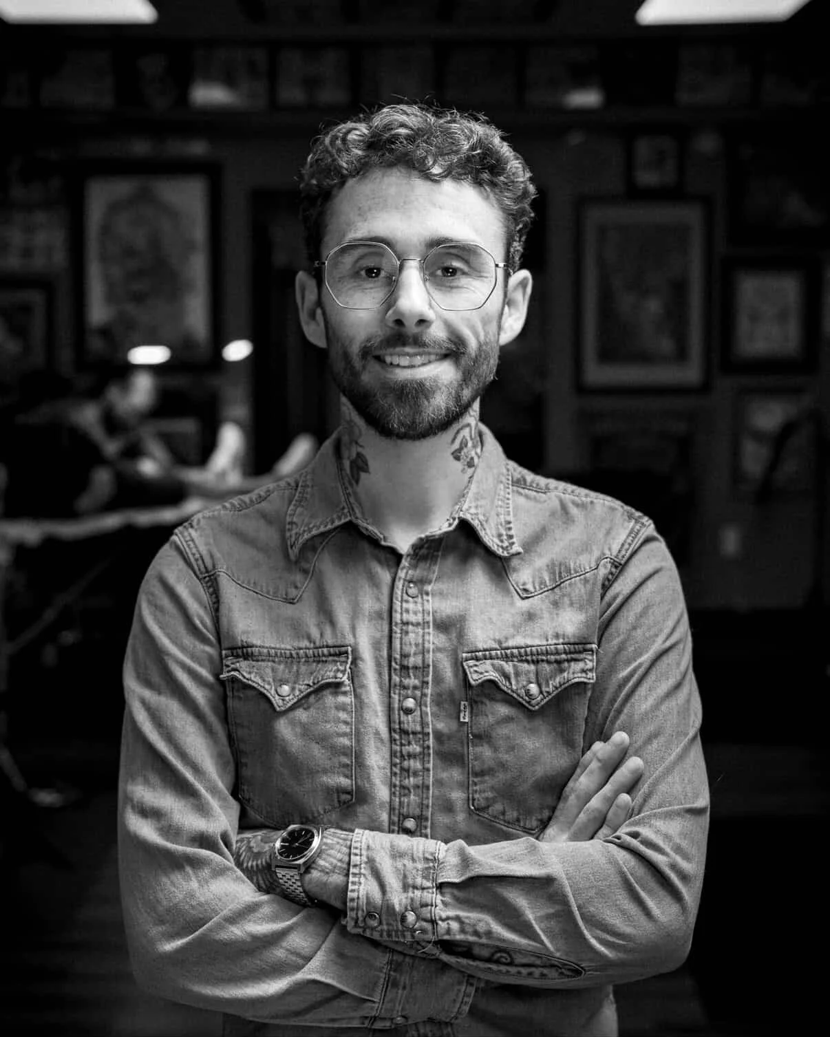 Black and white photo of a young man with curly hair, glasses, a beard, and tattoos on his neck. He is wearing a denim shirt with his arms crossed, standing in front of a blurred background with framed pictures. Evan Rice, Tattoo Artist.