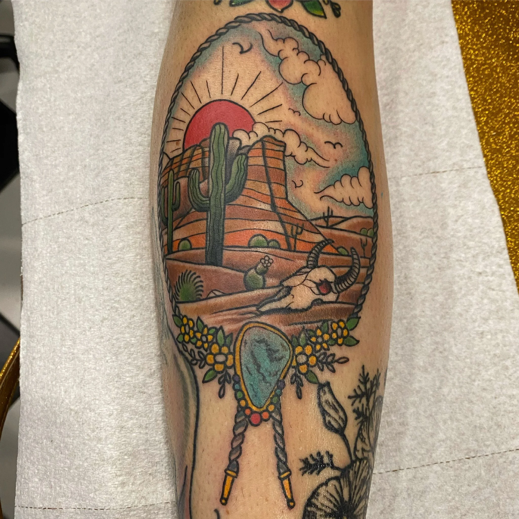 A colorful tattoo featuring a desert landscape with a red sun, clouds, cacti, and a mountain with lines. There's a longhorn skull with exaggerated horns at the bottom, surrounded by yellow flowers and green leaves, and a turquoise gemstone hanging fr