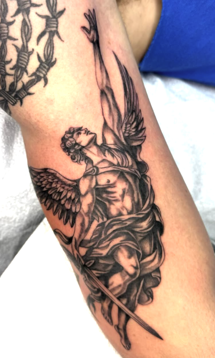 Black and gray tattoo of a winged, muscular figure holding a spear, with detailed shading and a dynamic pose, on person's arm.