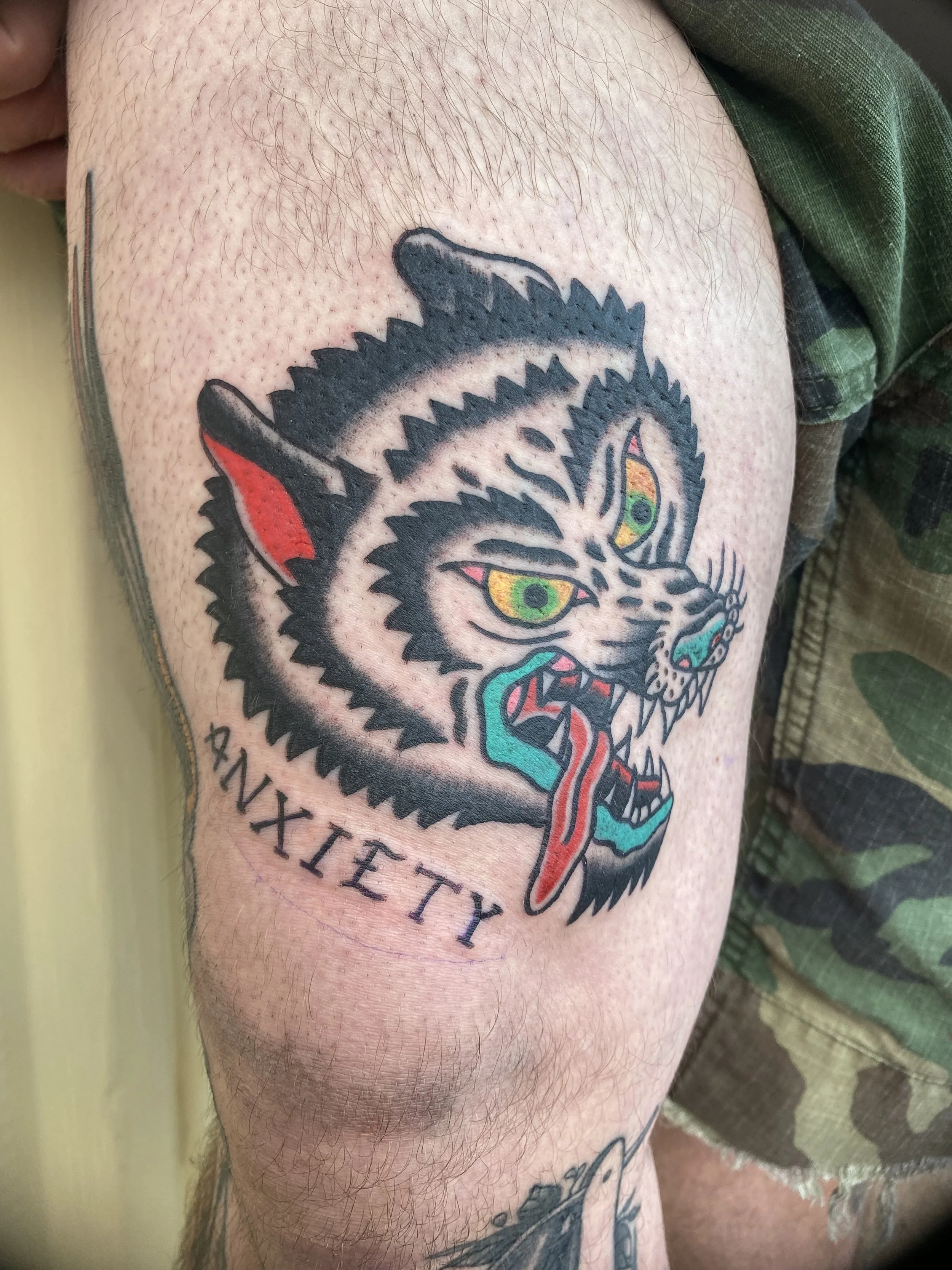 Tattoo of a fierce tiger's head with green eyes, showing sharp teeth, tongue out, and black and gray stripes, with red in the ear and tongue, and the word 'ANXIETY' below.
