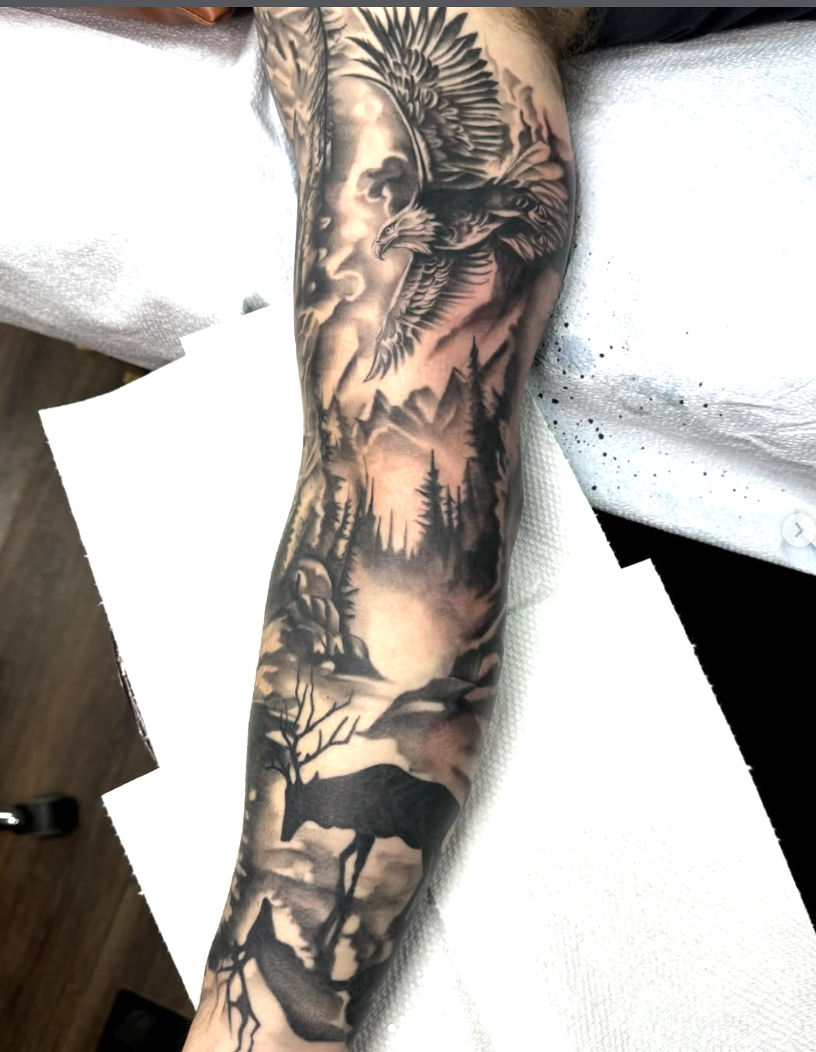Tattoo of a hunting scene on a person's arm, featuring a moose, pine trees, mountains, an eagle, and a cloudy sky.