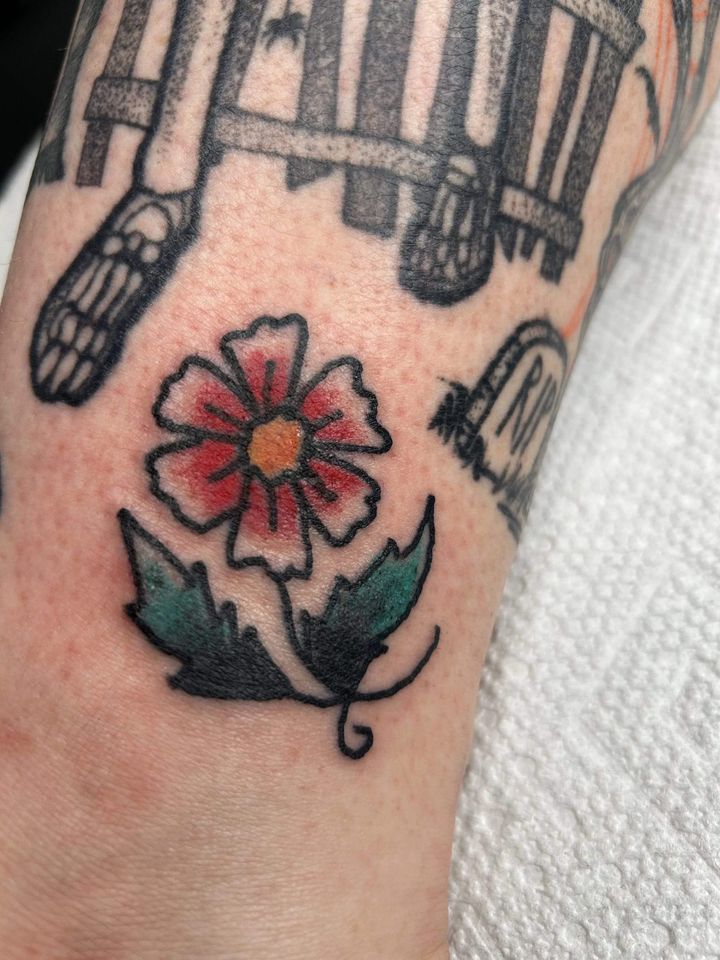Colorful tattoo of a flower with red petals and a yellow center, and green leaves on a person's arm.