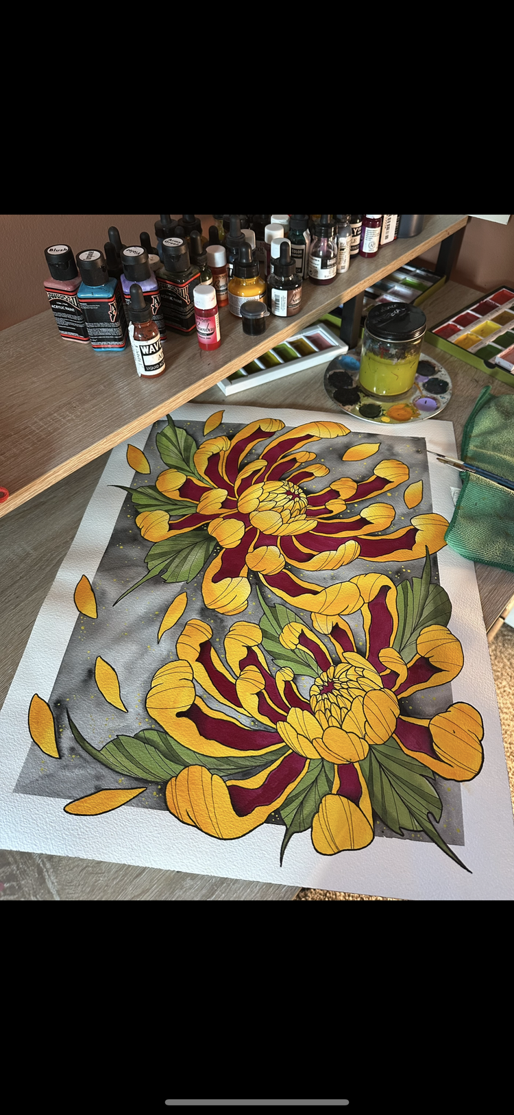 Colorful hand-painted floral artwork on a white paper, featuring yellow and maroon flowers with green leaves and falling yellow petals, on a wooden desk with art supplies and paint bottles.