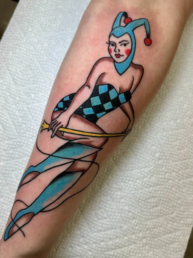 Tattoo of a woman with a jester hat, checkered stockings, holding a yellow wand, and wearing blue boots.