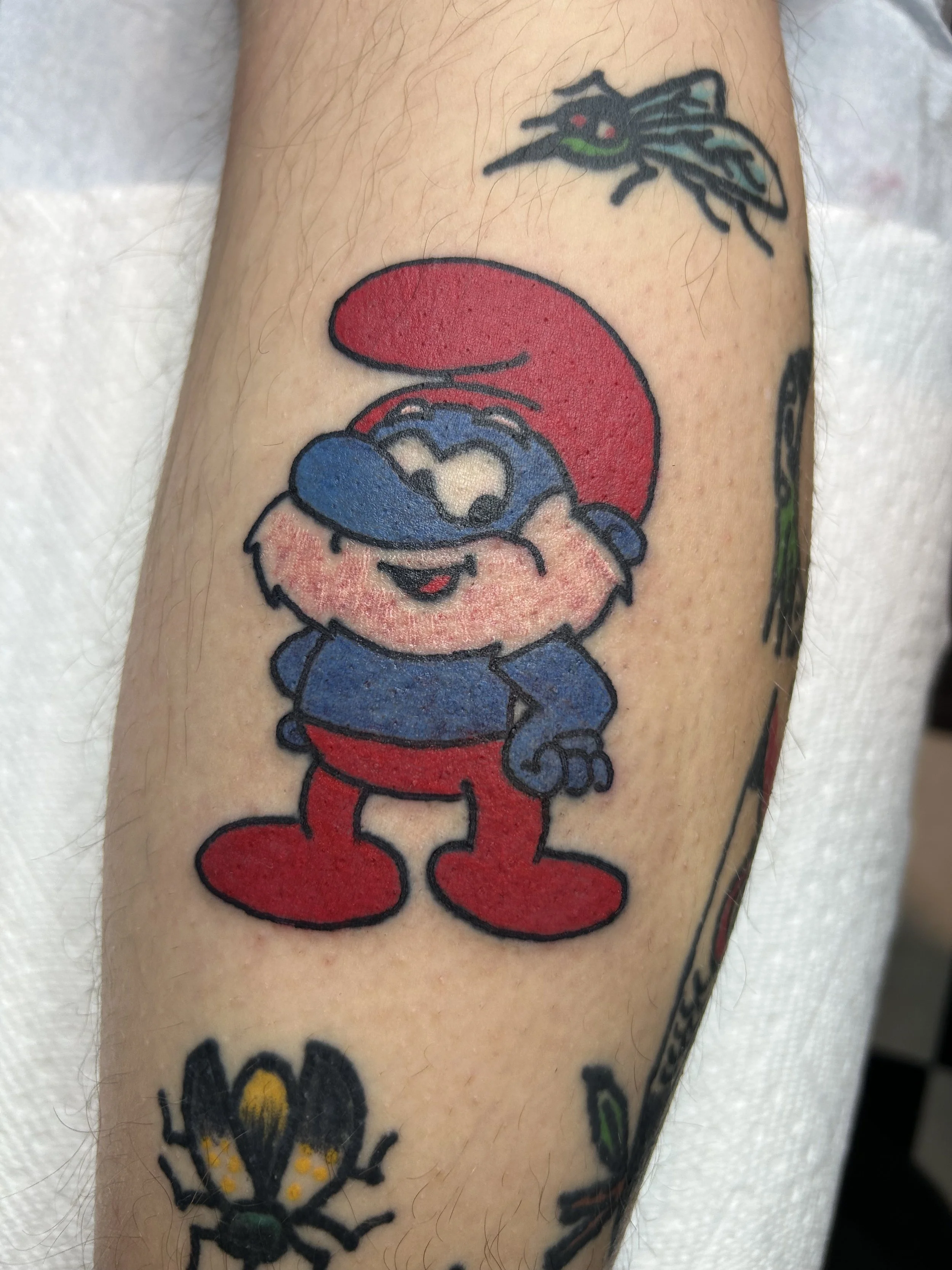 Colorful tattoo of Smurf wearing a red hat and red pants, smiling and standing with hands on hips, on person's arm.
