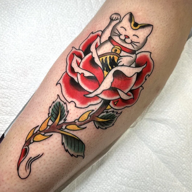 Tattoo of a Japanese lucky cat sitting inside a red rose on a person's forearm.