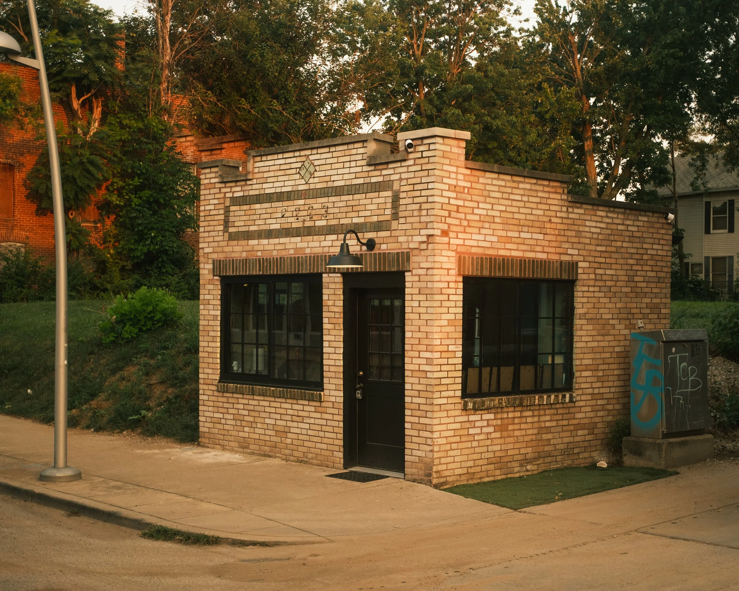 Small brick building with large windows, black door, and exterior light fixture, situated on sidewalk with graffiti-covered utility box to the right and hills with trees behind.