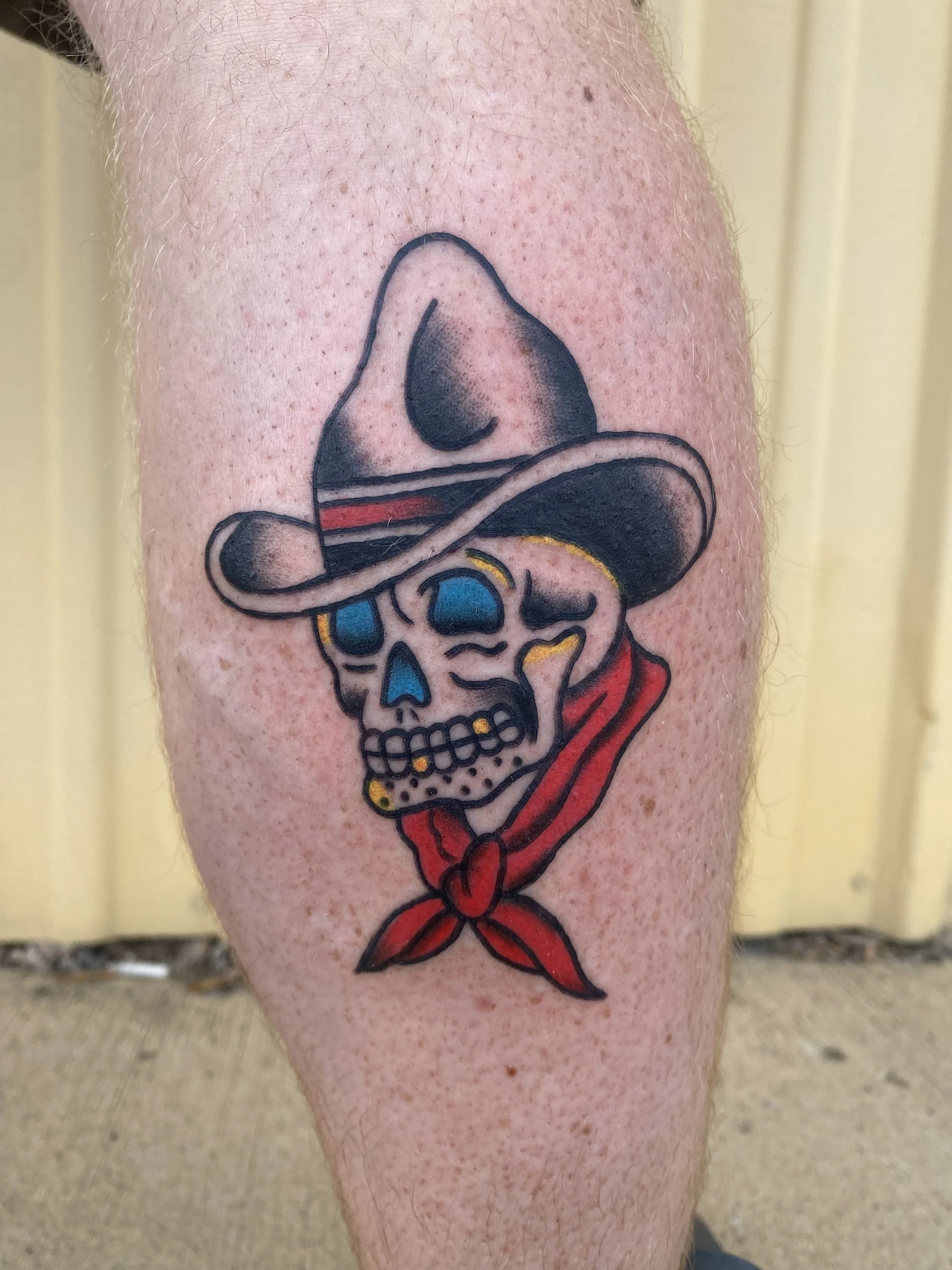 Tattoo of a skull wearing a cowboy hat and bandana, with blue eye sockets and gold teeth.