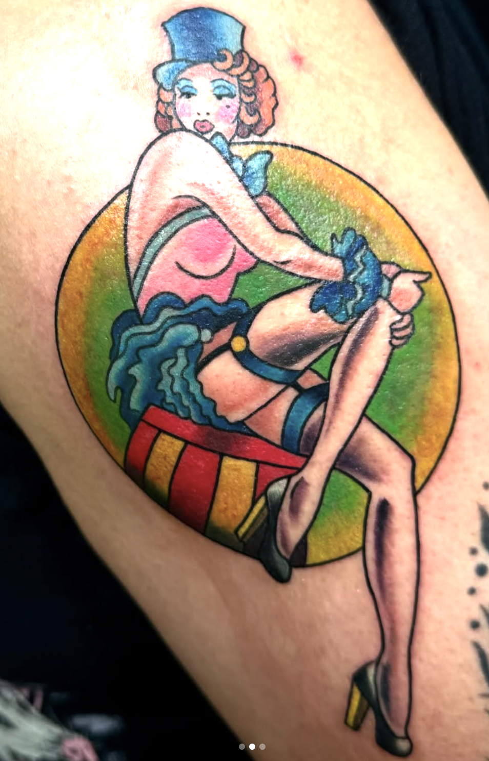 Colorful tattoo of a cabaret-style woman with curly hair, wearing a blue top hat, gloves, and fishnet stockings, sitting on a circus drum with a rainbow circle background.