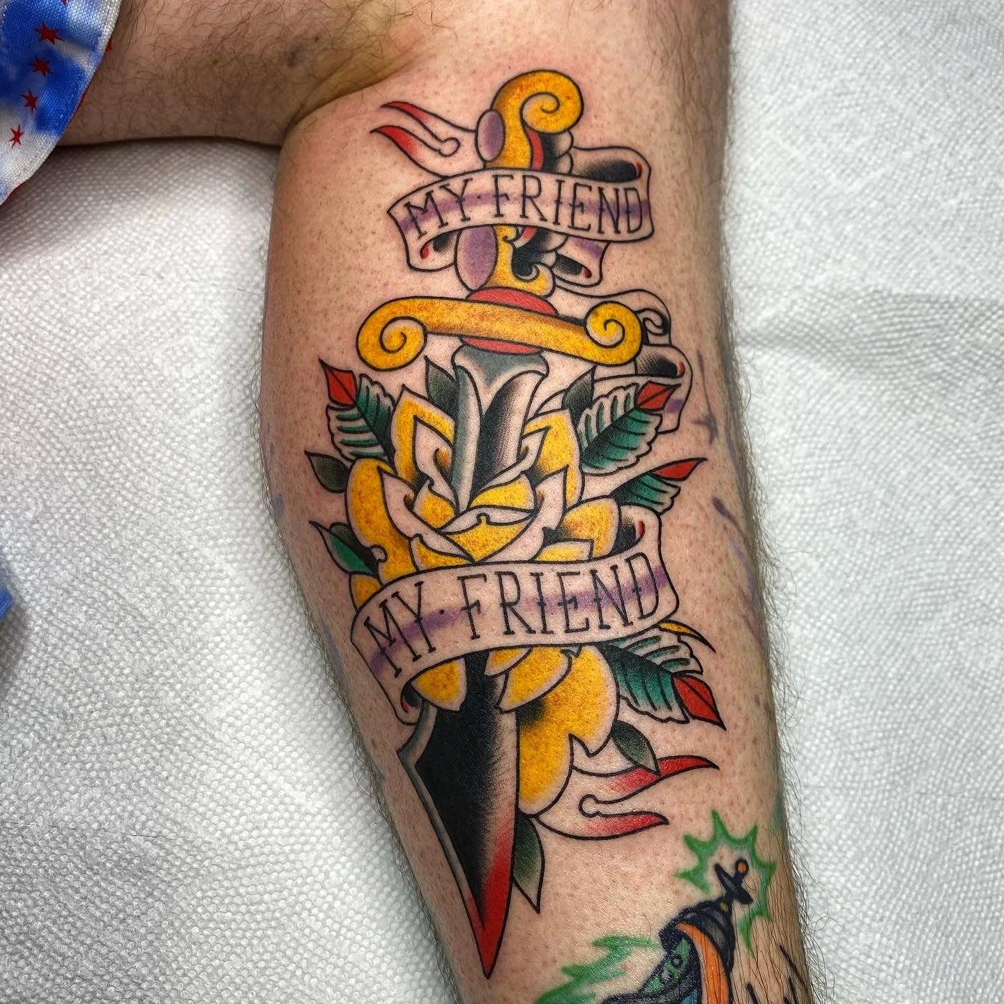 Colorful tattoo of a dagger with a rose and leaves, with the words 'My Friend' on banners at the top and bottom.
