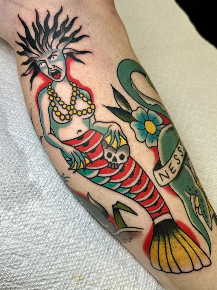 Traditional style tattoo of a mermaid with a fierce face, evil eyes, and wild hair, holding a skull with a yellow flower and a banner reading 'Ness'.