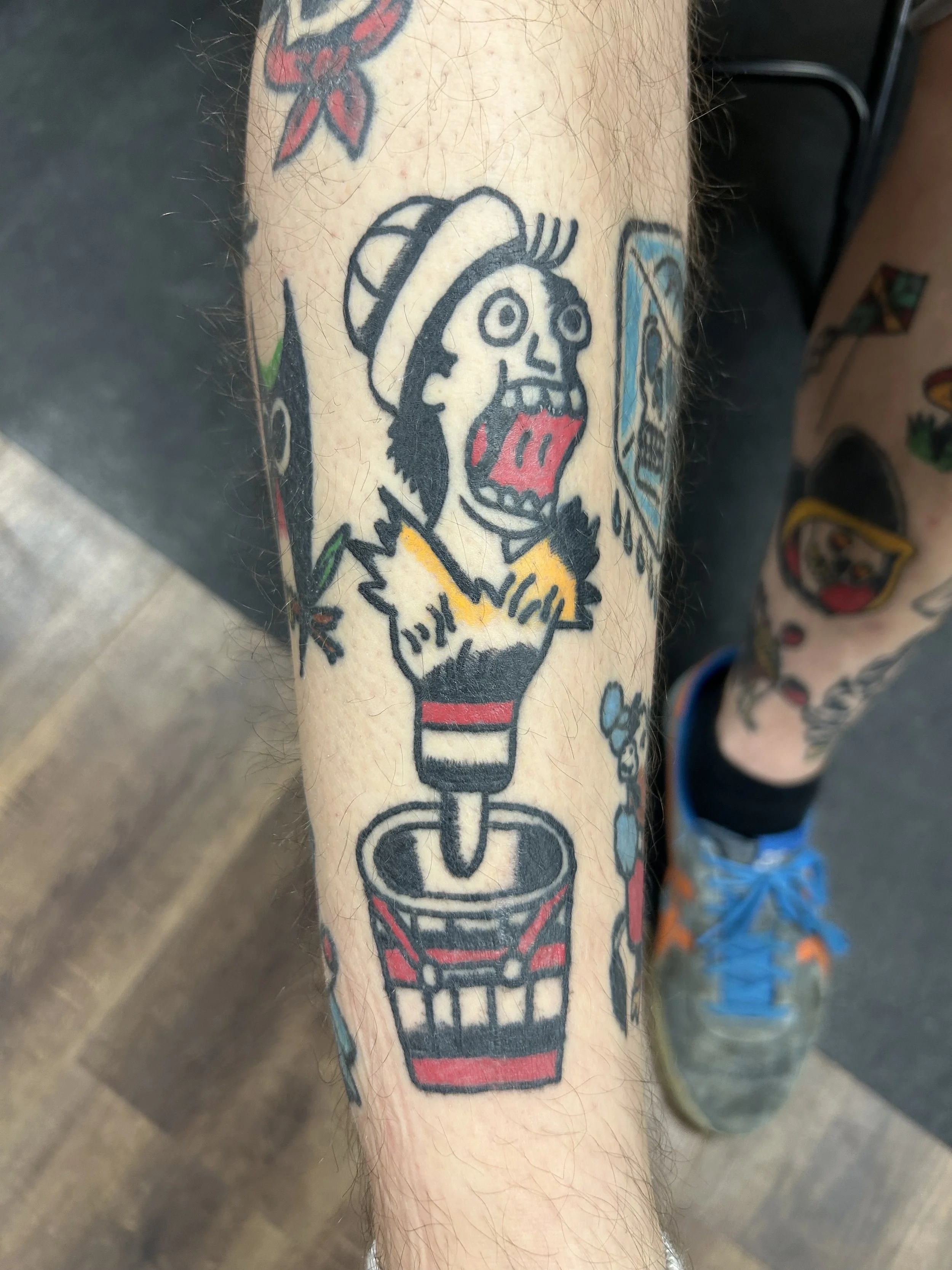 Tattoo of a cartoon character with a hat, shouting, with a lit firecracker in a soda cup, on the person's forearm.