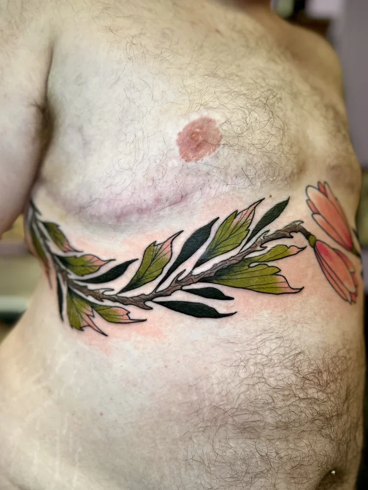 Close-up of a person's chest with a tattoo of a branch with green leaves and pink flowers around the nipple.