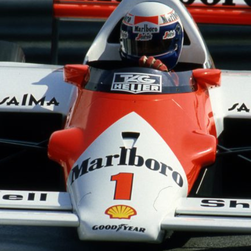 A race car driver sitting in a classic Formula 1 car with Marlboro branding, the number 1 on the front, and other sponsor logos, wearing a helmet.