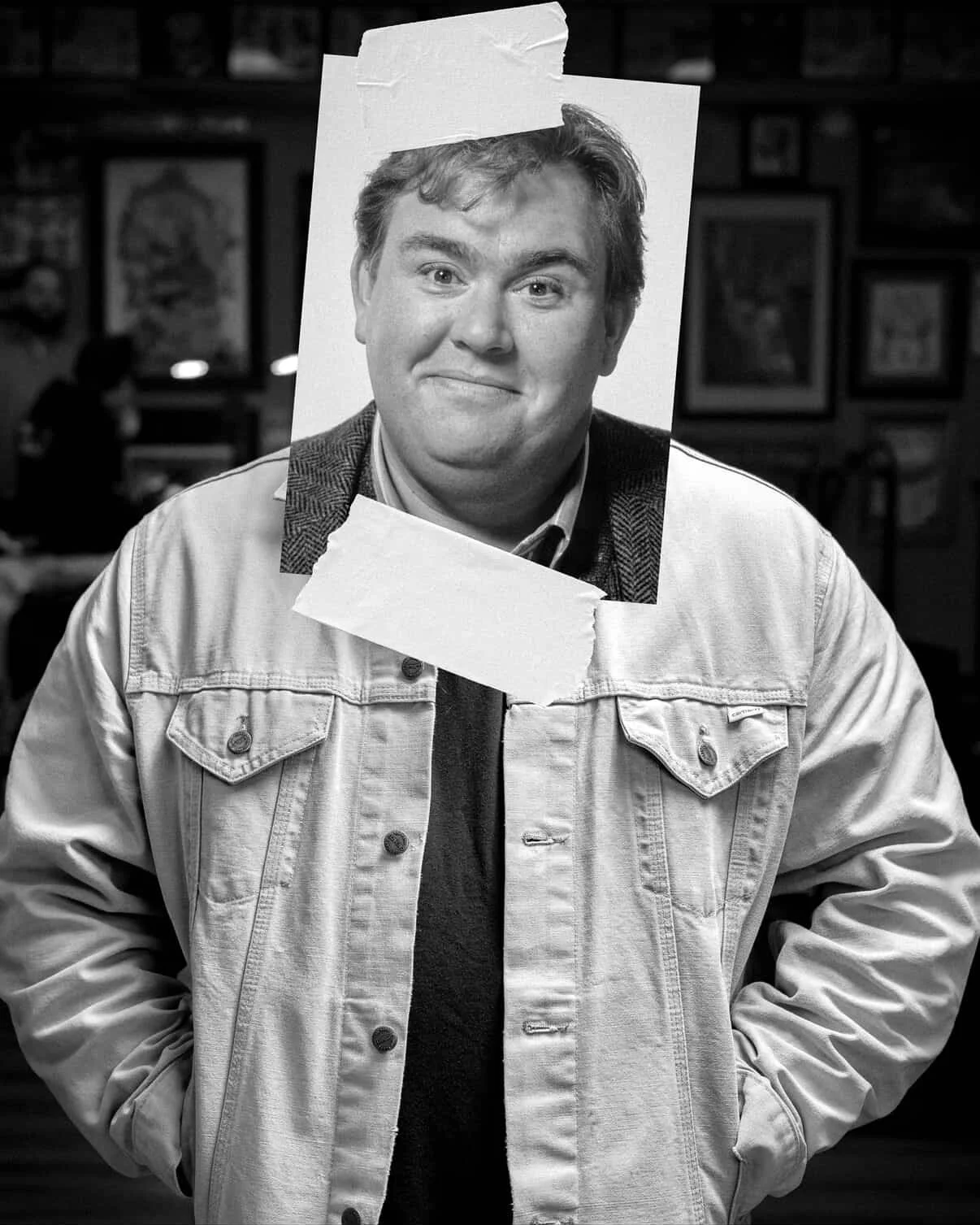 Black and white image of a man wearing a denim jacket with a newspaper-style photo of a smiling man taped to his face, with framed pictures on the wall in the background - Tom Tuttle, tattoo artist.