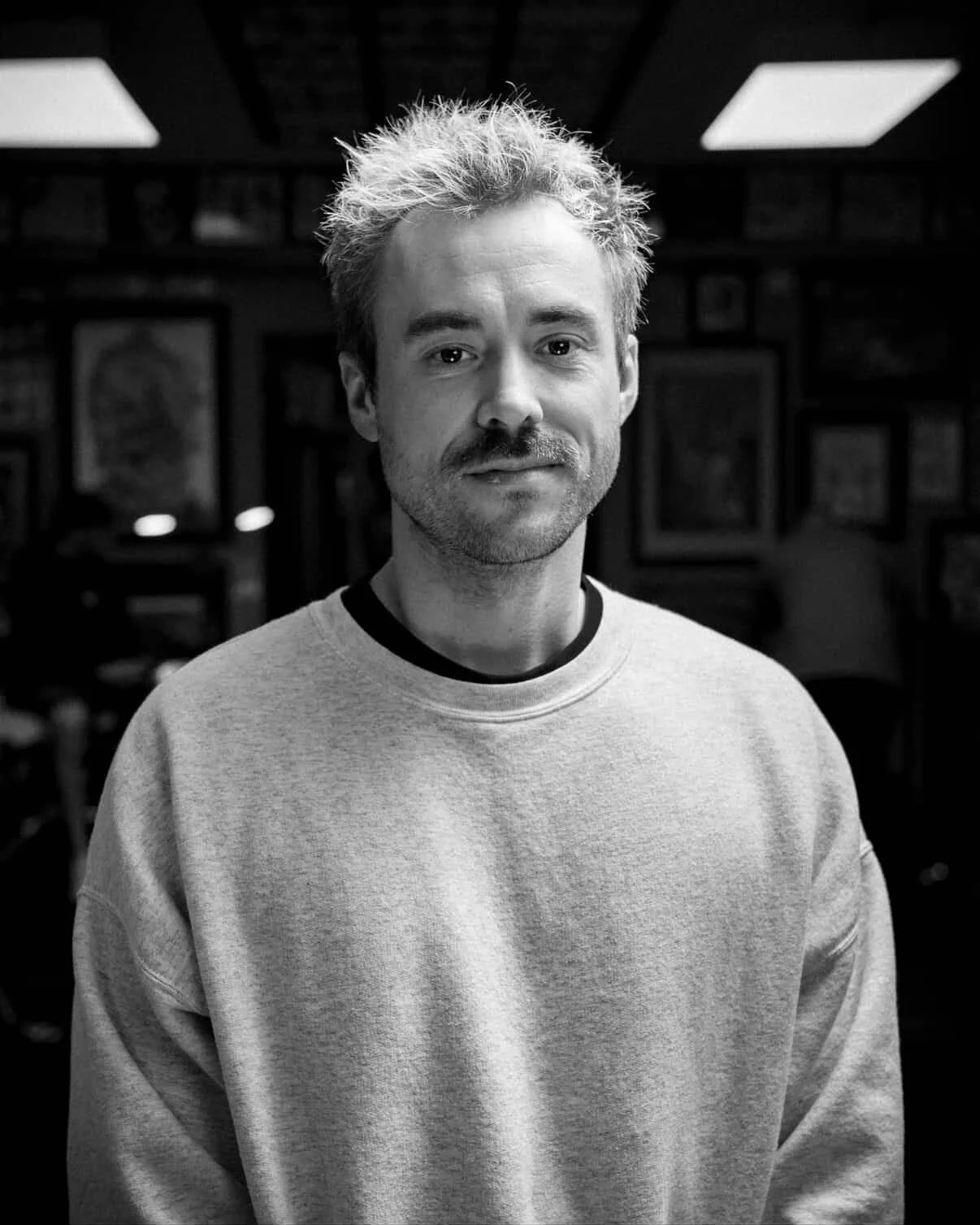 A black and white portrait of a man with light, spiky hair, a mustache, and a beard, wearing a light sweatshirt in an indoor setting with framed pictures in the background - Dylan Anderson, Tattoo Artist.