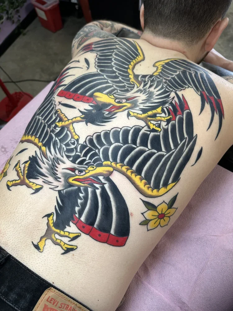 Tattoo of a fierce eagle with outstretched wings, clutching a banner that says "DUCE" in red letters, and a yellow flower on the lower side of the back.