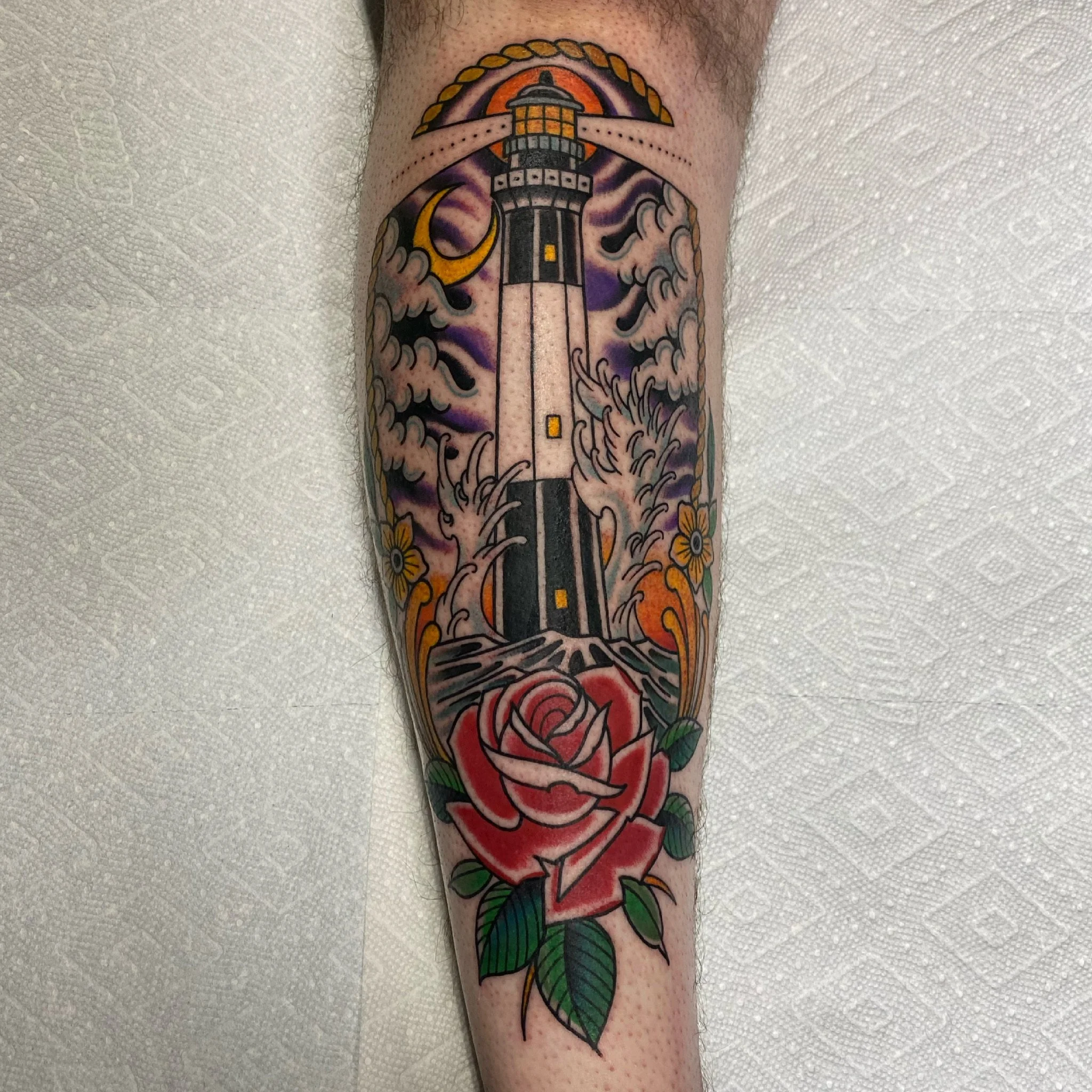 Tattoo of a lighthouse with waves, moon, and clouds above, and a red rose with green leaves below.