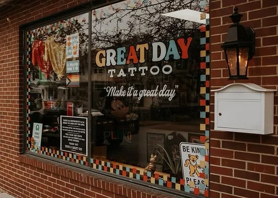 Storefront window with colorful lettering that reads 'Great Day Tattoo' and the tagline 'Make it a great day.' The window display includes tattoo designs and decorations, with a white mailbox and a black outdoor lantern mounted on the brick wall beside it.