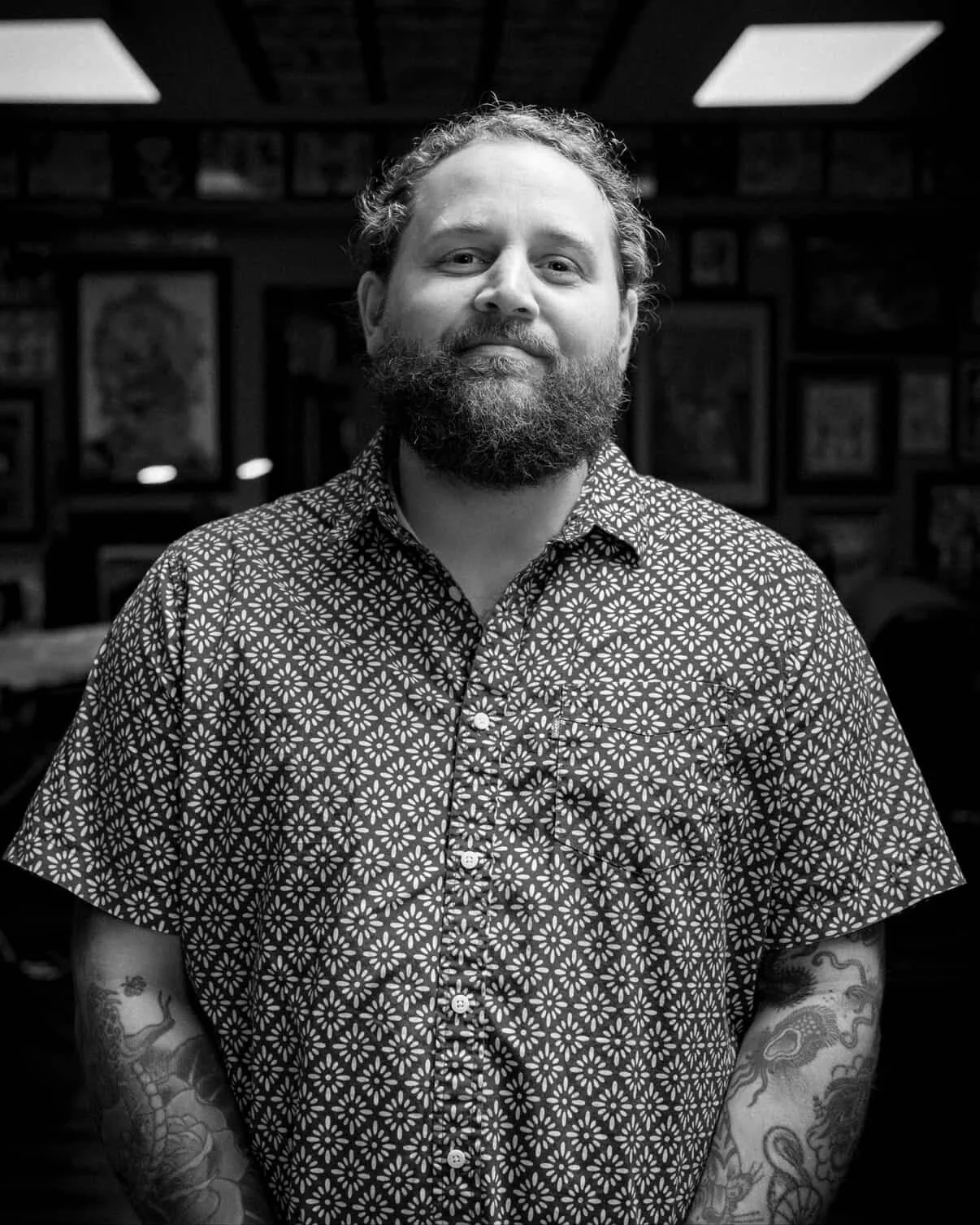 A man with a beard and tattoos on his arms, wearing a patterned short-sleeve button-up shirt, standing indoors with framed pictures on the wall in the background - Dustin Leach, tattoo artist.