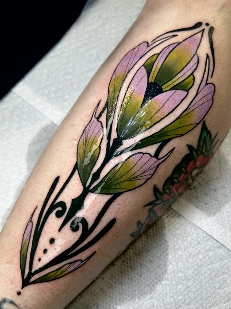 Colorful tattoo of a flower with pink and green petals, outlined in black on person's arm.