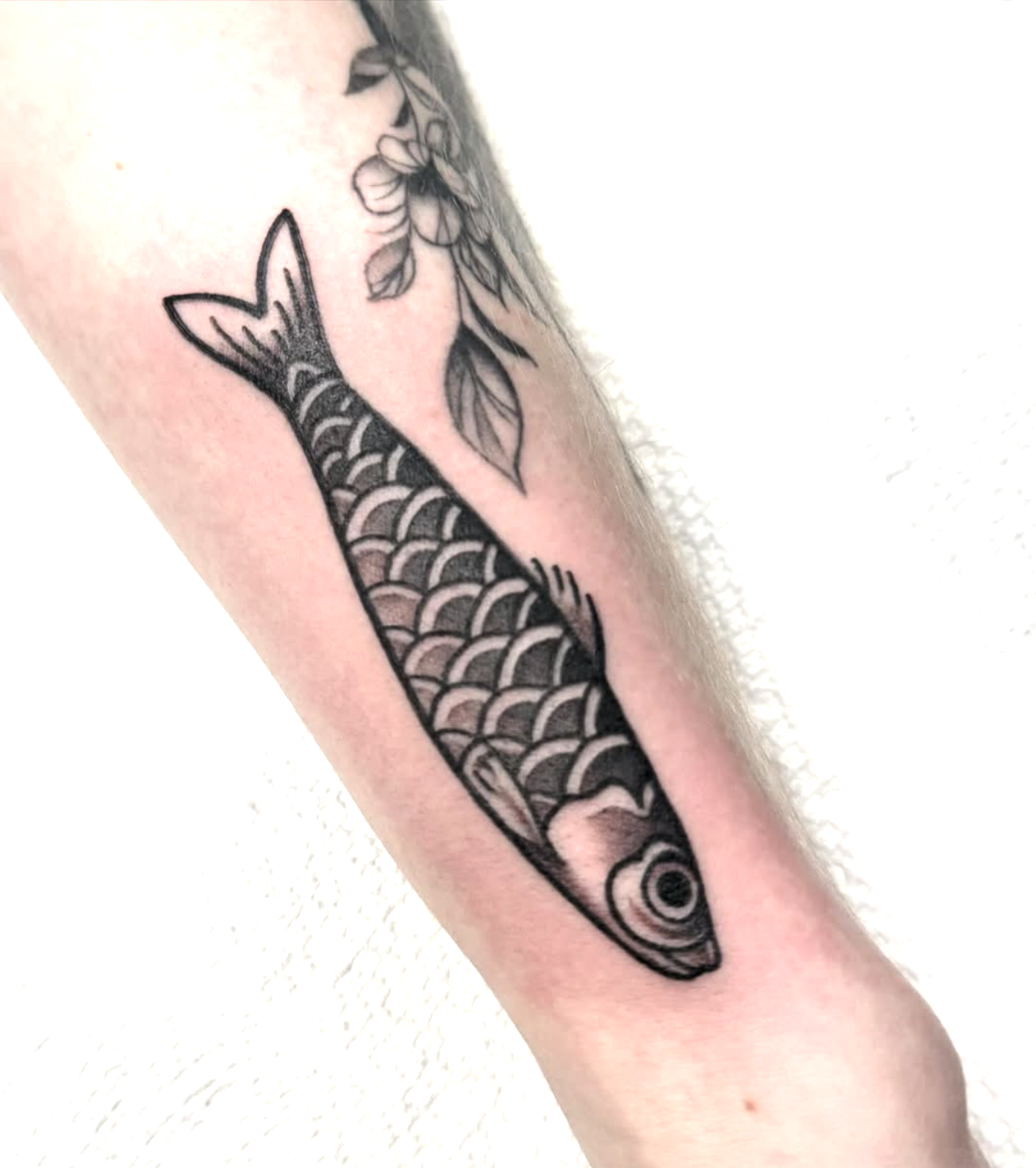 Tattoo of a fish with detailed scales on a person's arm, next to floral designs.