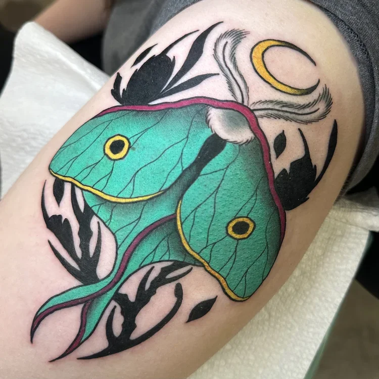 Colorful tattoo of a large green moth with yellow and black eyespots, surrounded by black silhouette branches, a yellow crescent moon, and wispy clouds.