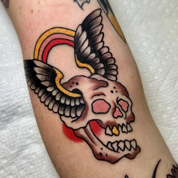 Tattoo of a skull with large wings, a rainbow arching over it, and a small yellow detail in the mouth.