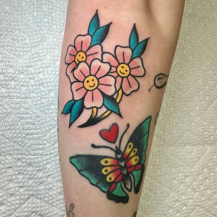 Colorful tattoo featuring pink flowers with yellow smiley faces at the center, green and blue leaves, a butterfly with yellow and red accents, and a small red heart.