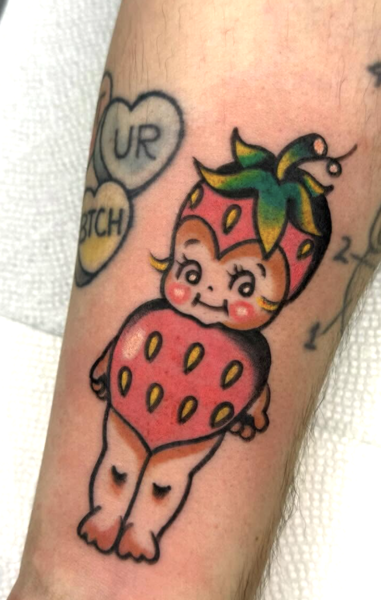 Colorful tattoo of a cute, winking child wearing a strawberry costume with green leaves on the head and a red body with yellow seeds, standing on feet with closed eyes.