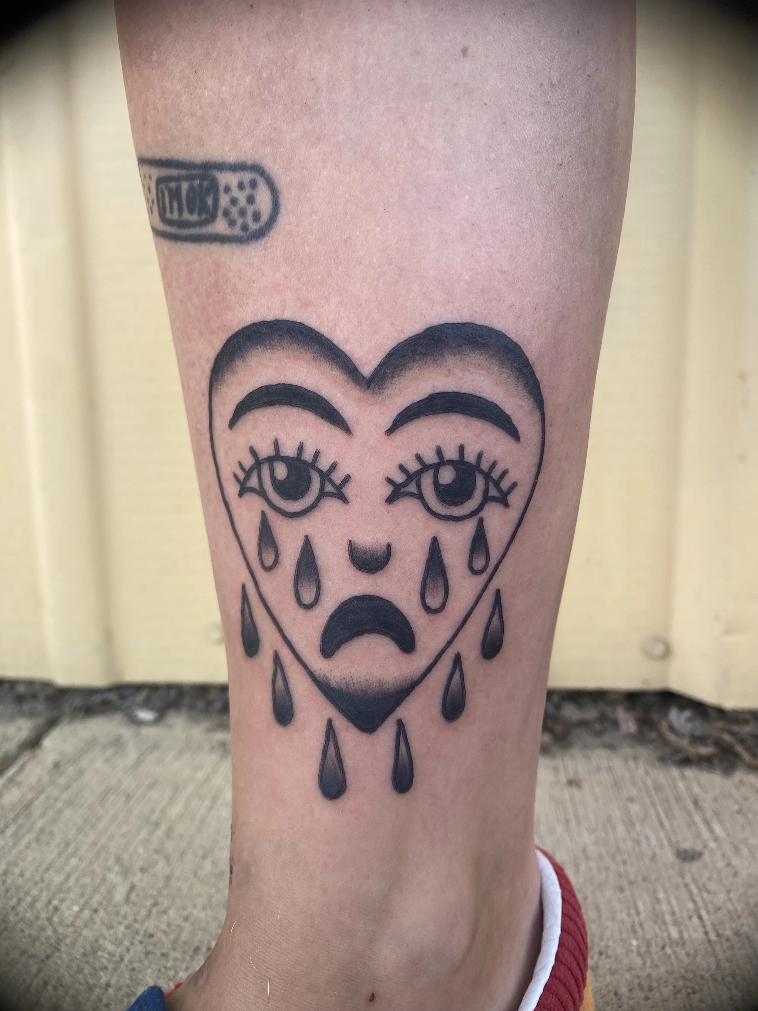 Tattoo of a sad face with teardrops inside a heart shape on a person's leg.
