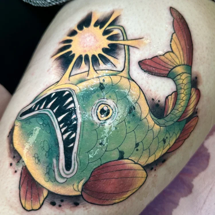 Colorful tattoo of a fish with an exaggerated open mouth and a light green body, red fins, and a large eye, with a sunburst and rays above its head.