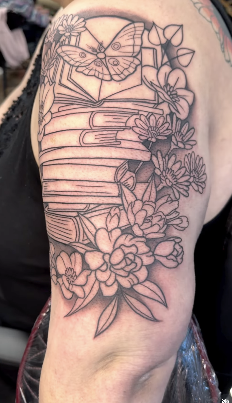 Black outline tattoo of a butterfly sitting on a stack of books, surrounded by flowers and leaves.