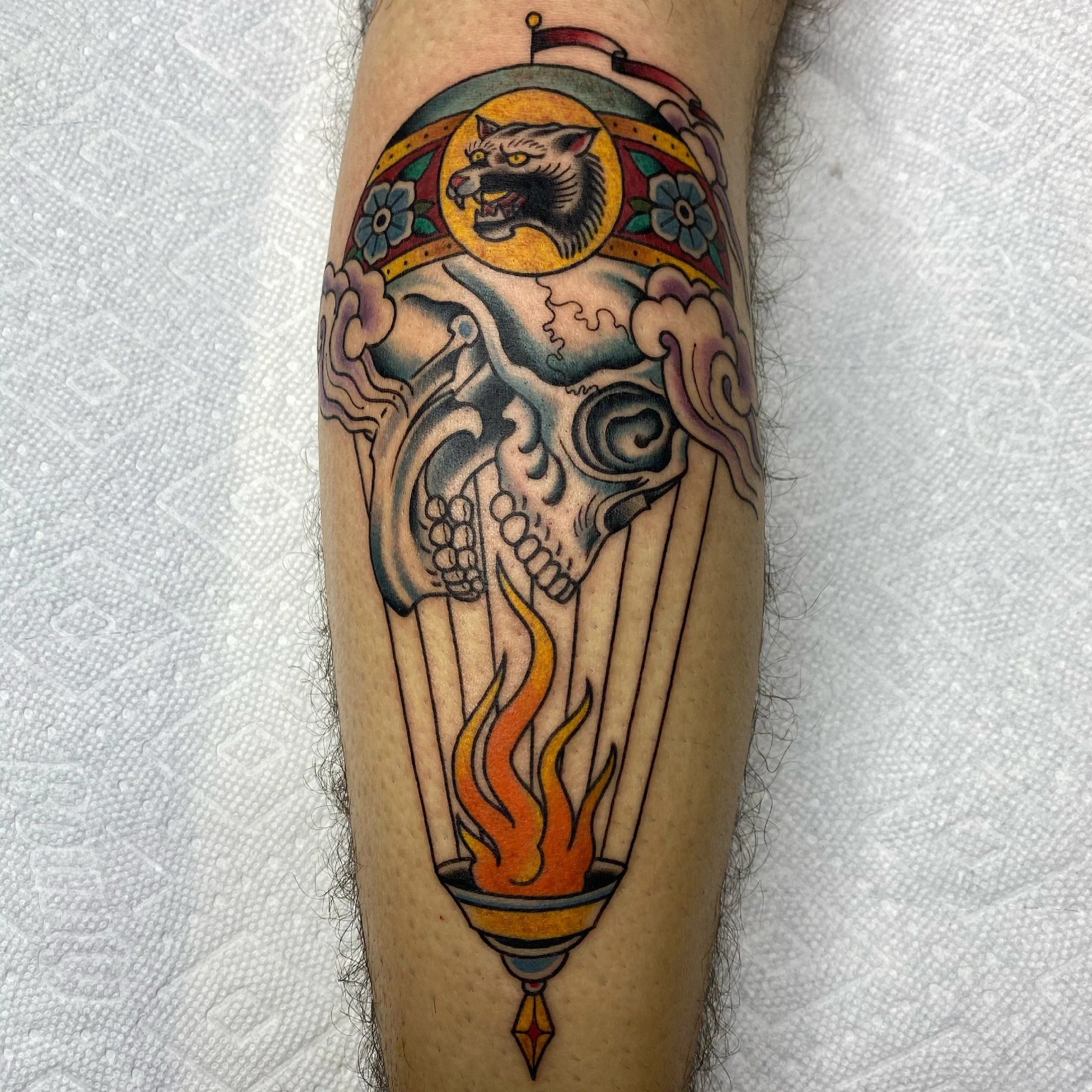 Tattoo of a skull with a flaming bowl beneath it, a cat face with a crown above, and decorative clouds and flowers.