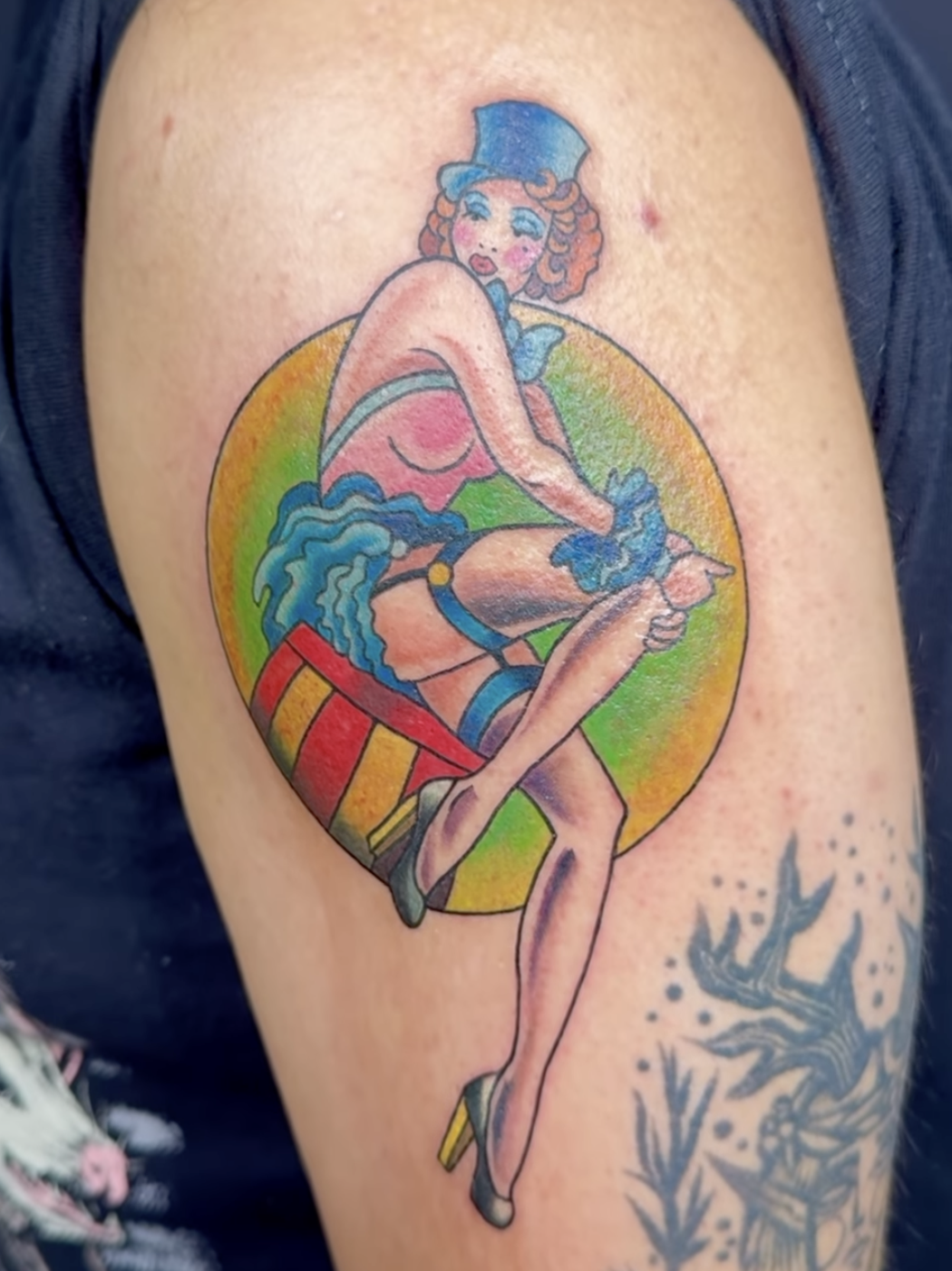 Colorful tattoo of a burlesque dancer with curly hair, wearing a top hat, gloves, and high heels, sitting on a striped stool with a rainbow background.