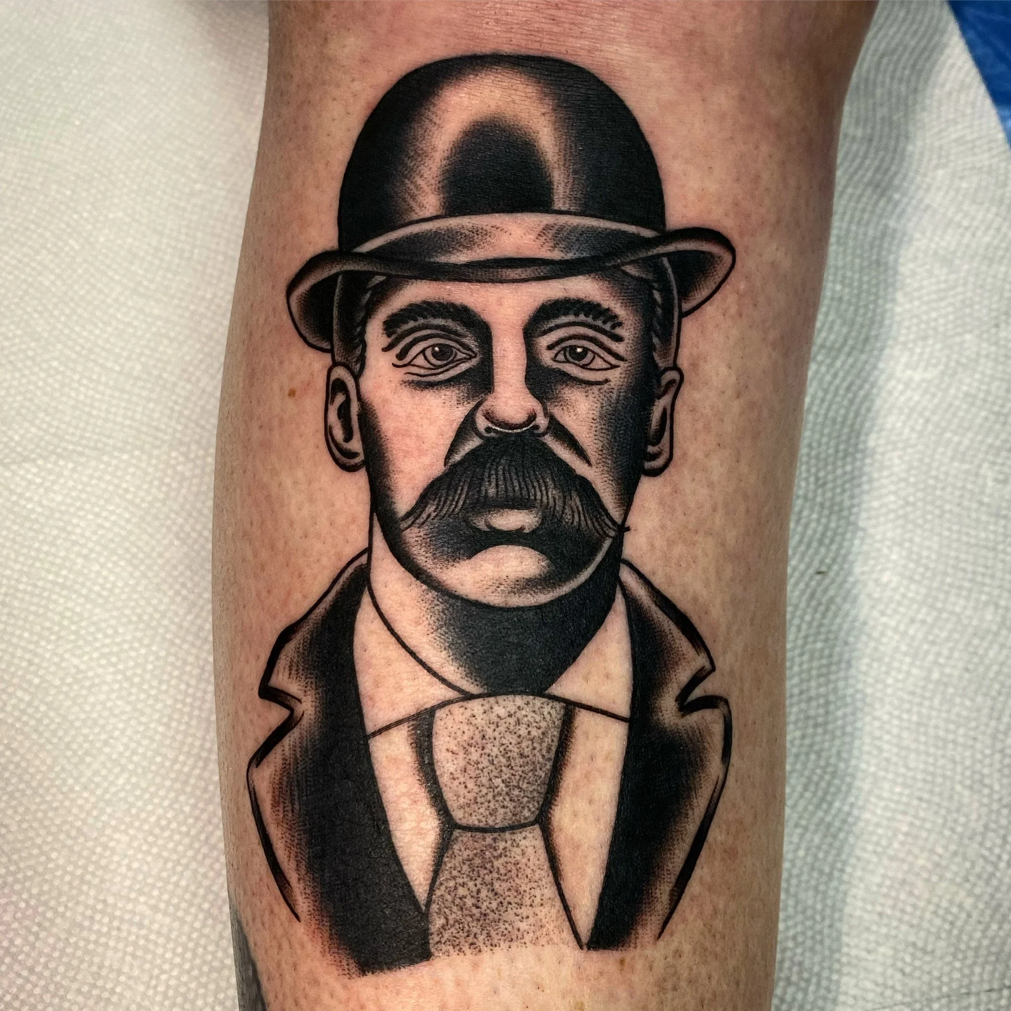 Black and grey tattoo of a man with a mustache, wearing a bowler hat, a suit, and a tie, on a person's arm.