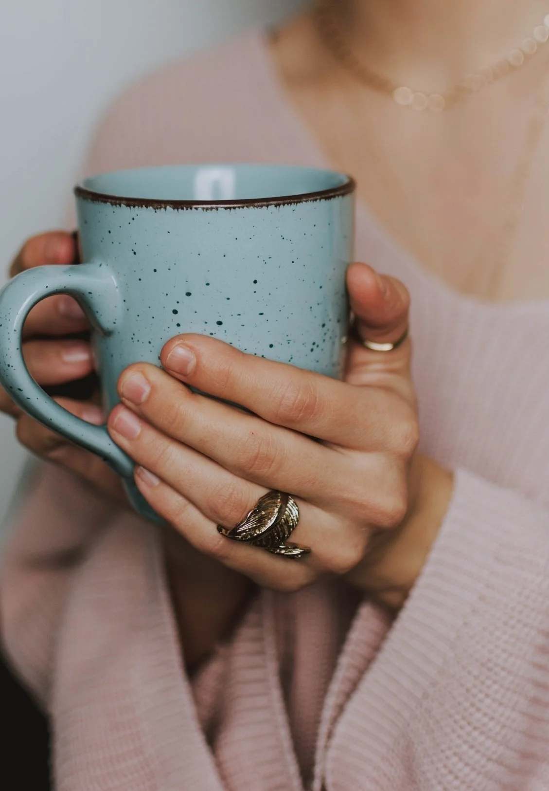 A person holding a speckled blue mug with both hands, wearing multiple rings and a pink sweater.