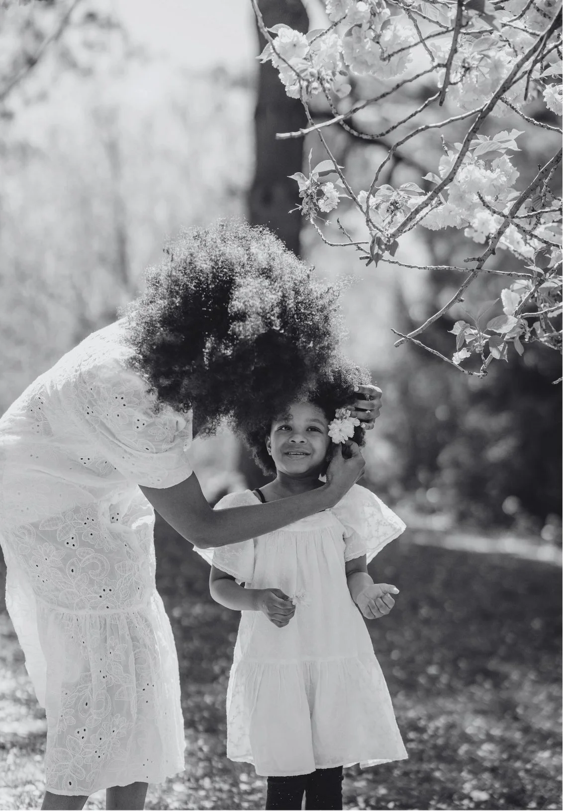 A woman with large curly hair gently touching a young girl’s face, both dressed in white, outdoors with trees and flowers, in black and white.