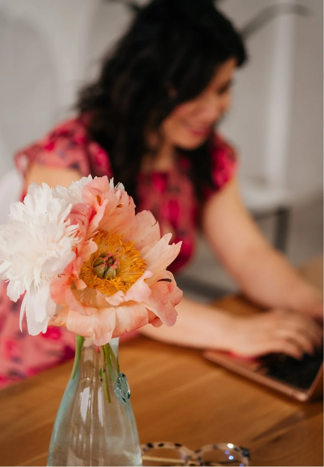 Close-up of a pink and white peony flower in a glass vase on a wooden table with a woman in a pink floral dress working on a laptop in the blurred background.