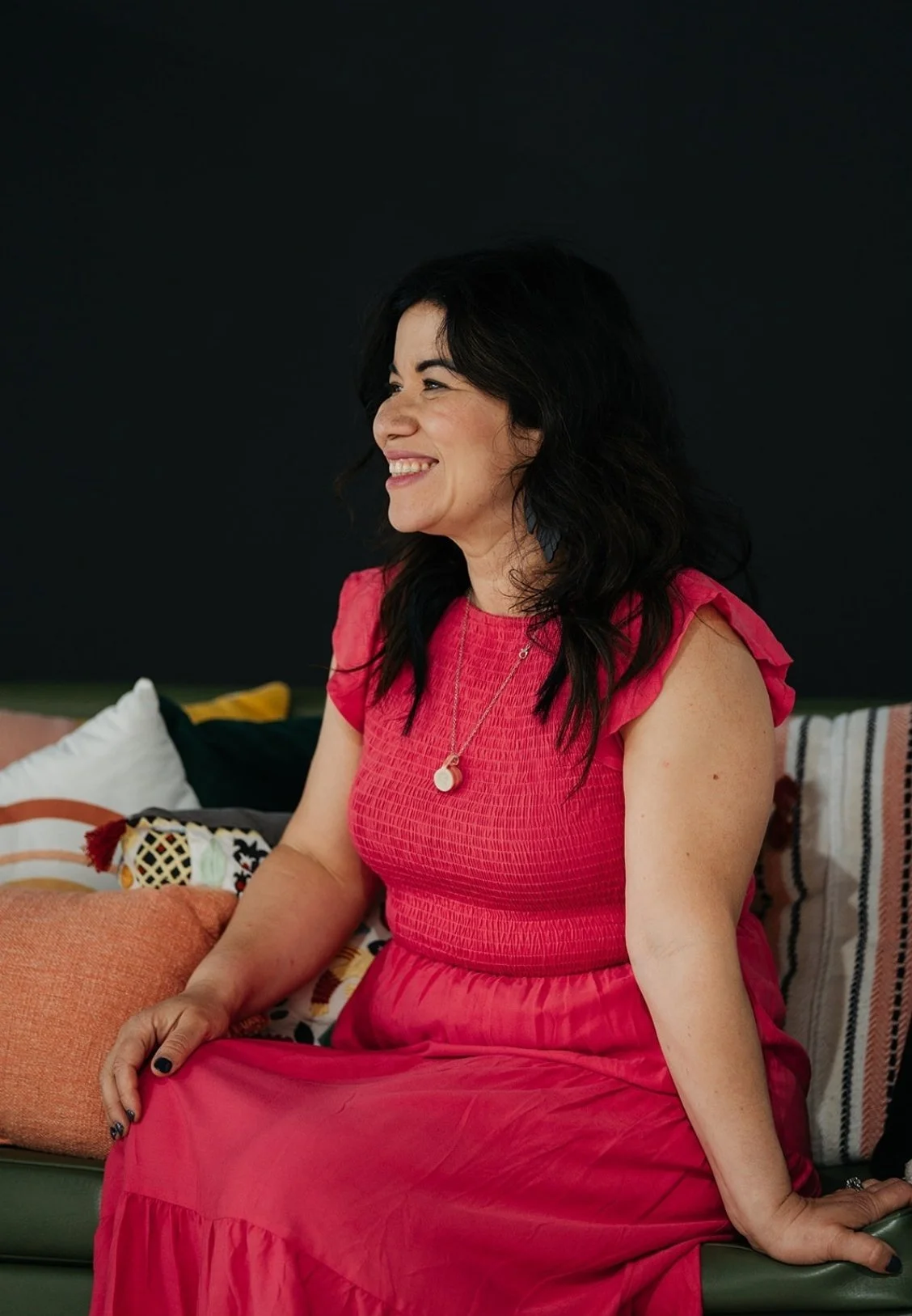 Adaris Pickett, Boston-based therapist, sitting on a couch smiling and looking to her left. The background is dark, and colorful cushions are on the couch.