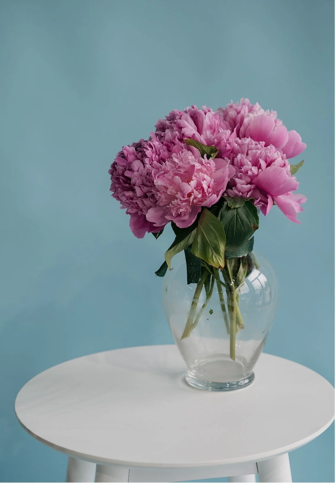 A bouquet of pink peonies in a clear glass vase on a white round table against a light blue background.